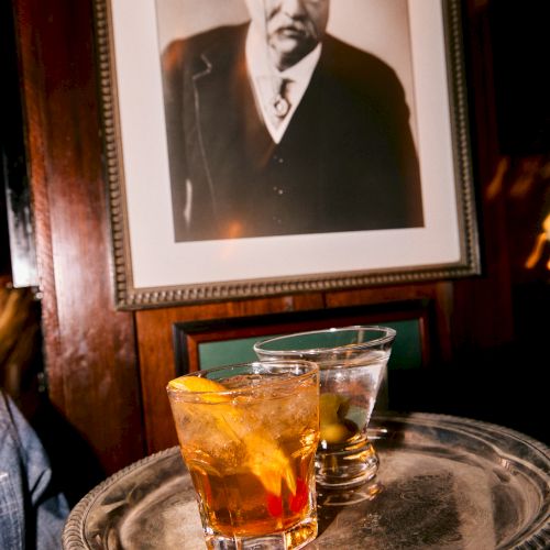 A framed black-and-white portrait hangs above a tray with two glasses of amber drink and ice.