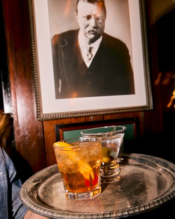 A framed black-and-white portrait hangs above a tray with two glasses of amber drink and ice.