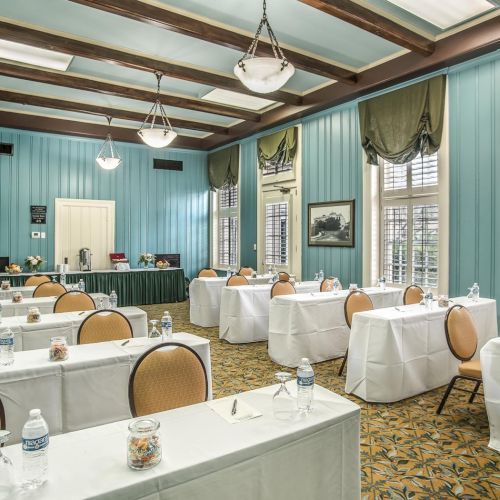 A conference room setup with turquoise walls, long tables covered in white linens, tan chairs, water bottles, and decorative lighting fixtures.