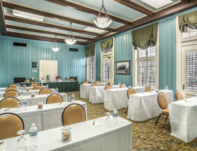 A conference room setup with turquoise walls, long tables covered in white linens, tan chairs, water bottles, and decorative lighting fixtures.