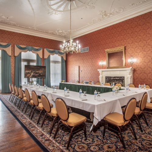A formal dining room with a long U-shaped table set for a meeting, white tablecloths, gold chairs, ornate red wallpaper, chandeliers, and a marble fireplace.