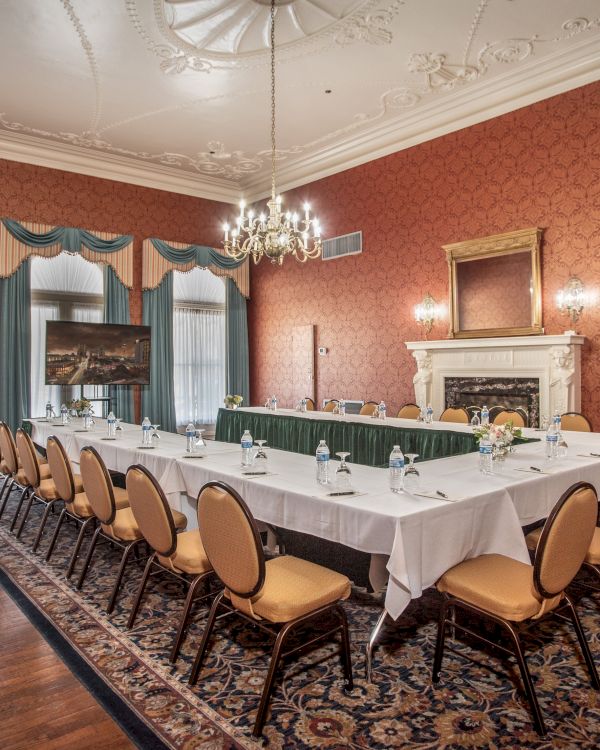 A formal conference room with a long U-shaped table, green chairs, chandelier, ornate red wallpaper, fireplace, and draped windows.