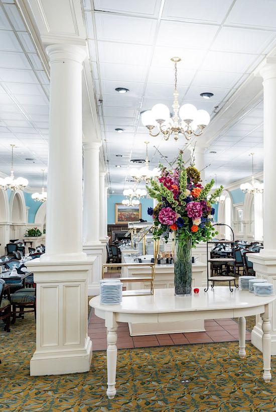 A bright, elegant restaurant with chandeliers, white columns, arched windows, blue walls, and a central floral display on a pale pink table.