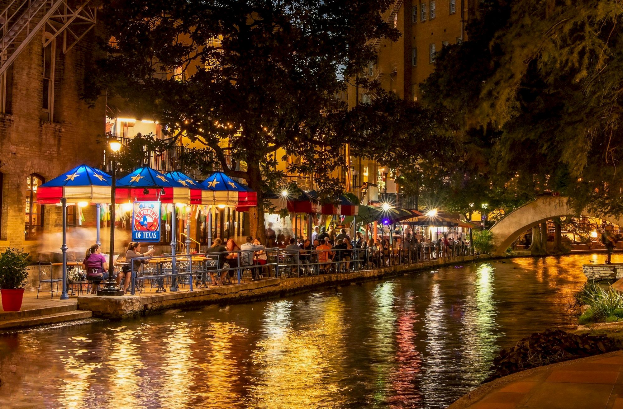 Night canal scene with a riverside patio, blue umbrellas, warm lights reflecting on water, people dining under trees by the waterway.