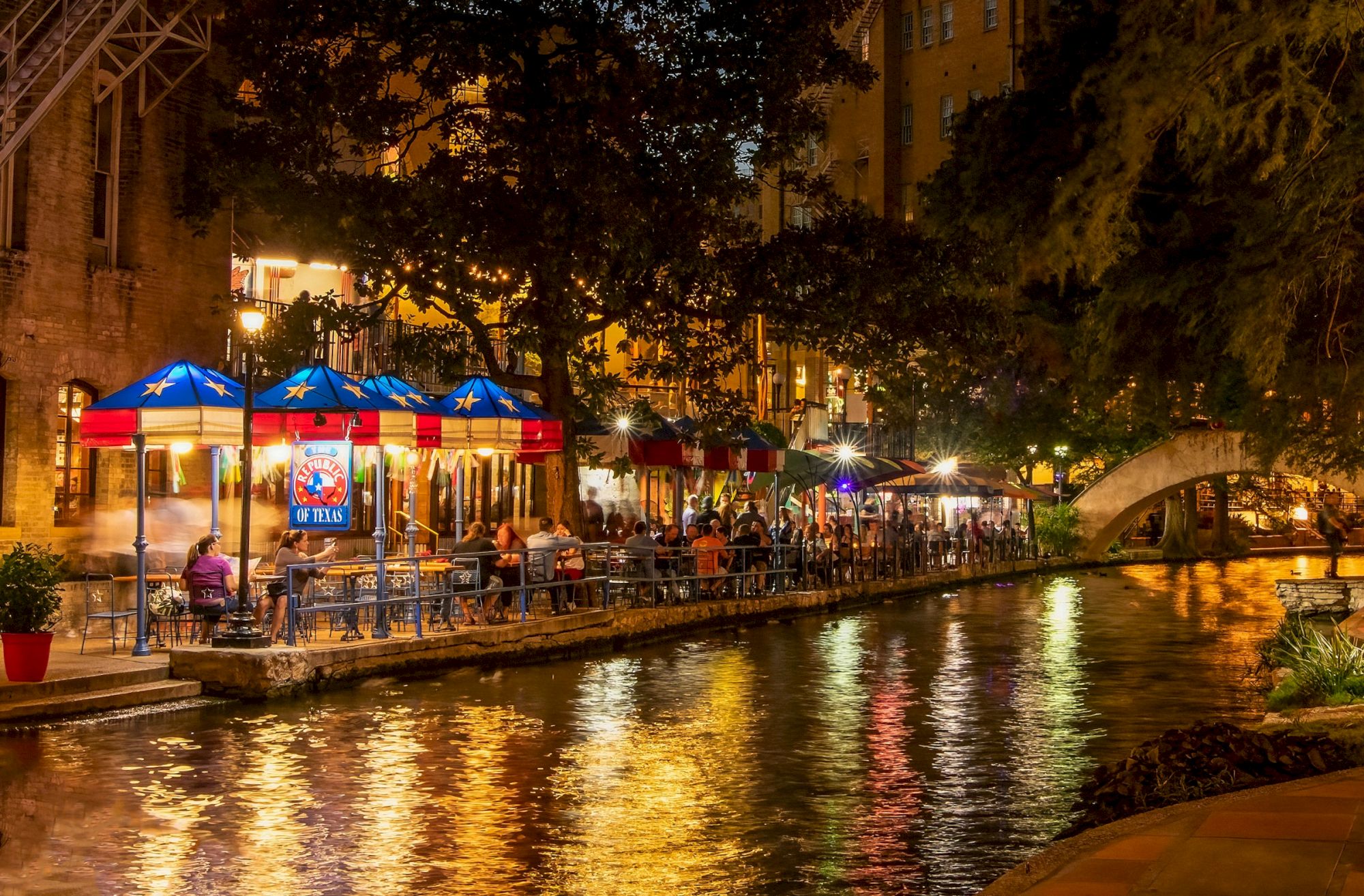 A lively riverside cafe scene at night with outdoor seating under blue umbrellas, colorful lights reflecting on calm water.