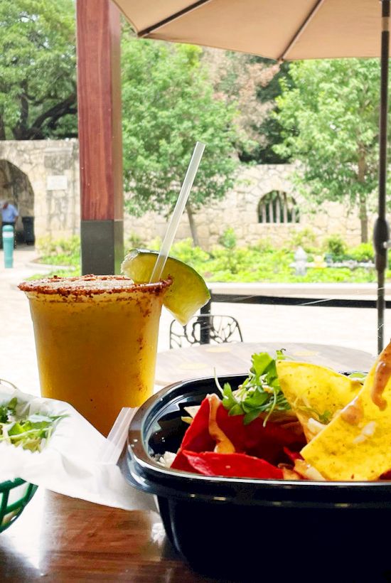 Outdoor cafe table with chips and dip, a salad, iced drink with straw, and an umbrella against a sunny courtyard backdrop.