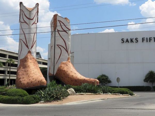 A pair of oversized cowboy boots serving as a sculptural entrance, placed in front of a Saks Fifth Avenue building with blue sky and clouds.