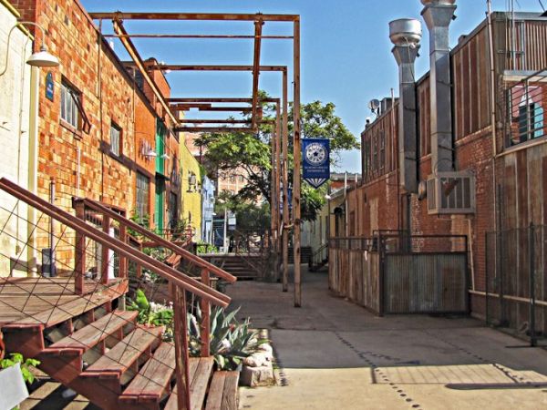A sunny alley with brick walls, stairs on the left, metal railings, potted plants, and a walkway lined by fences and lamps.
