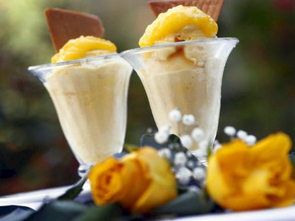 Two elegant vanilla desserts in small glass cups topped with pineapple and a chocolate wafer, set beside yellow roses.