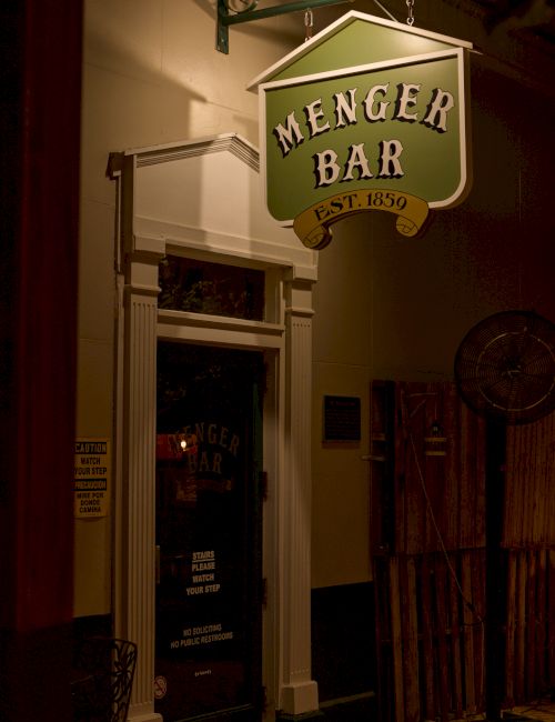 A green hanging sign reads &ldquo;MENGER BAR&rdquo; (est. 1859) inside a dim doorway.