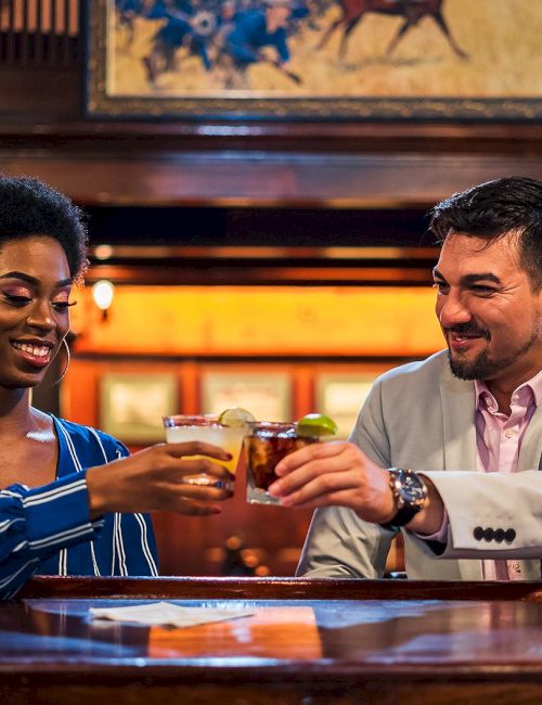Two people sharing a toast at a bar, smiling and clinking glasses, warm lighting and wood interior.