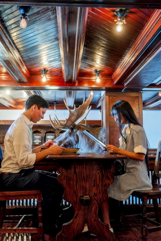 A couple sits at a bar, looking at a large mounted deer head between them, with warm wooden decor and ceiling lights overhead.