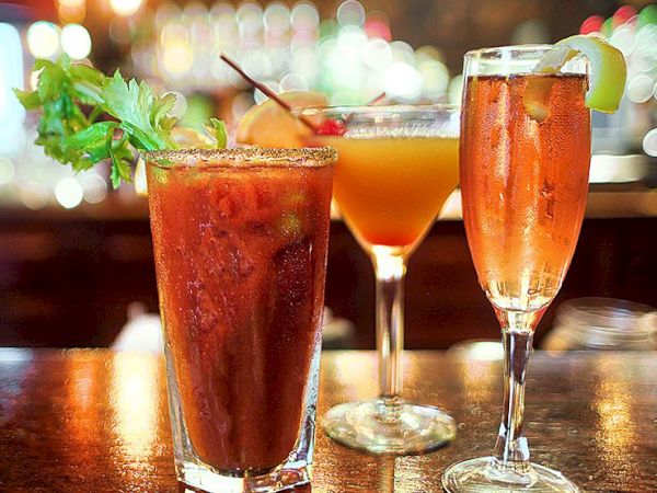 Three cocktails on a bar: a tall dark drink with a mint garnish, a peachy-orange martini, and a sparkling wine with a lime wedge, all lit warmly.