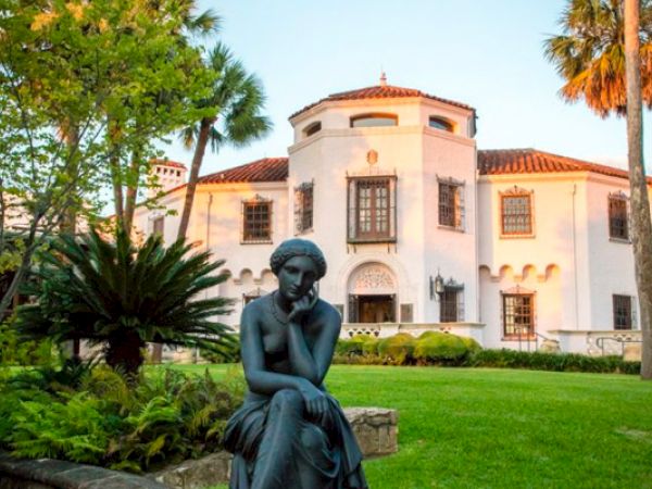 A statue of a thoughtful seated figure in front of a white Mediterranean-style villa with palm trees and well-kept lawn, sunny day.