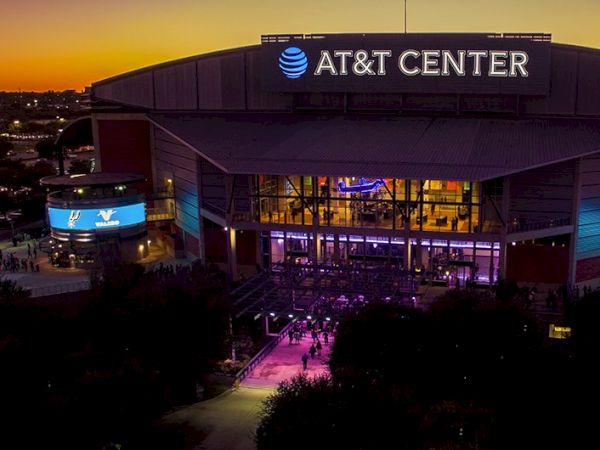 Aerial view of AT&T Center at dusk, illuminated entrance with purple carpet, crowd gathering outside, city lights in the background.