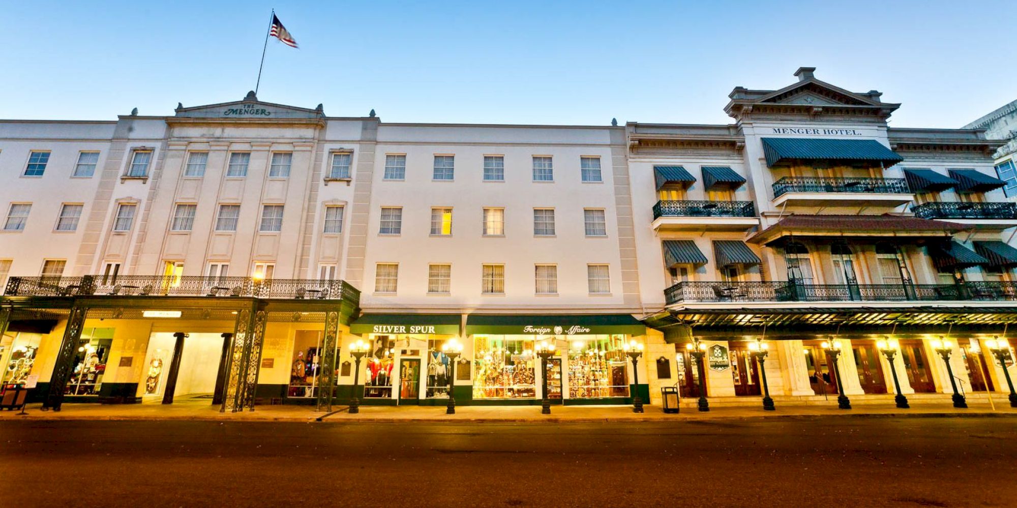 A row of stylish, well-lit storefronts with a white multi-story building on the left and a traditional building with balconies on the right; American flag on top.
