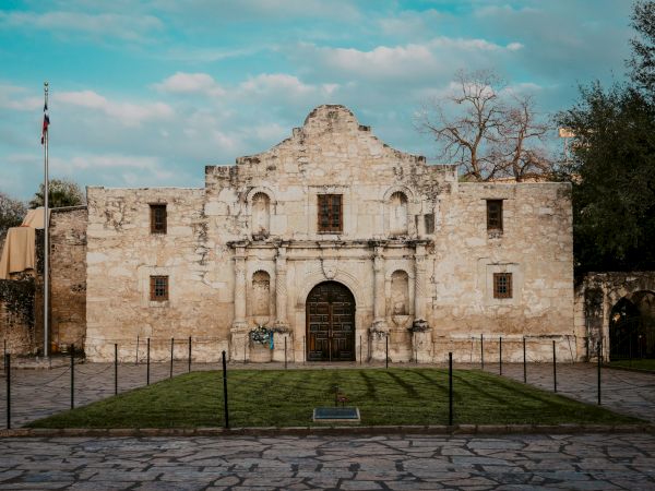 An old stone ruin with a central arched doorway, flanked by windows, a flagpole on the left, and a small lawn fenced off in front, clear sky.