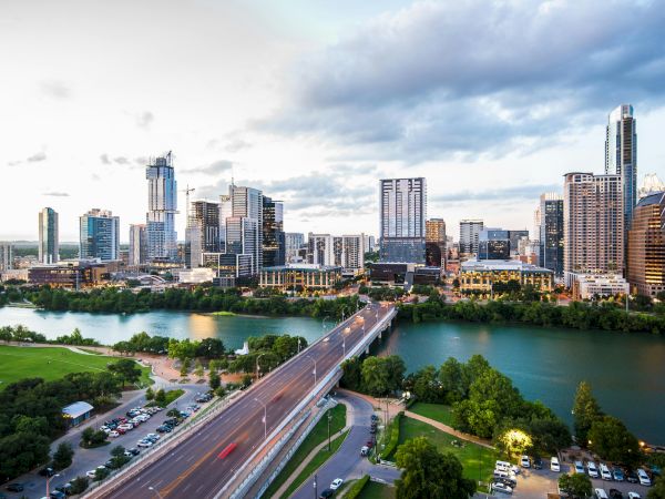Acity skyline across a river with a bridge, tall buildings, green parks, and a vibrant downtown under a partly cloudy sky.