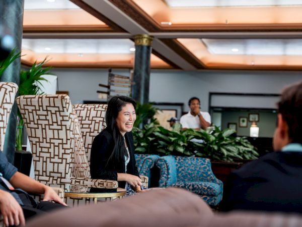 A woman smiles while seated in a stylish lounge with patterned chairs; others relax in the background of a bright hotel lobby.