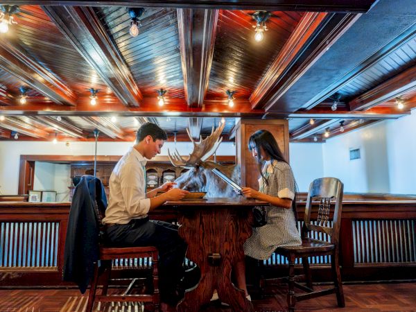 Two people sit at a rustic bar table, facing a mounted deer head on the wall while chatting and sipping drinks in a cozy, wood-paneled pub.