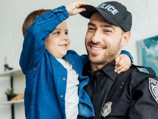 A smiling police officer in uniform holds a happy child, who salutes with a blue jacket, sharing a warm, friendly moment.