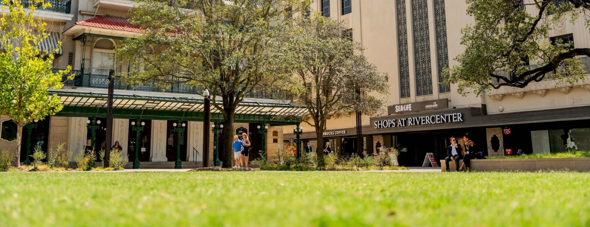 A sunny street with classic storefronts, a green lawn in the foreground, and people walking near the hotel and shops, around a tree-lined boulevard.