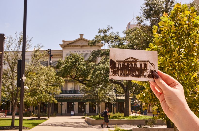 A hand holds a vintage postcard of the Menger Hotel, with the grand hotel building and leafy trees behind it.