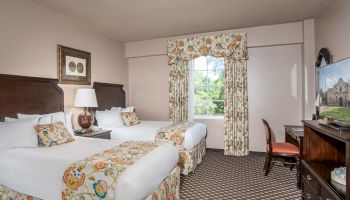Two queen beds with floral runners and pillows in a hotel room, a desk with chair, a window with floral curtains, and a dresser with a TV, all in a beige-carpeted room.