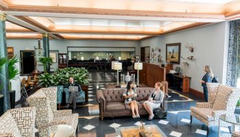A stylish hotel lobby with checkered floor, vintage chairs, a central couch area, plants, and a few guests relaxing or chatting.