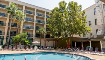 A hotel-style courtyard with a swimming pool, lounge chairs, palm trees, and a multi-story building surrounding the deck, sunny day.