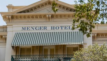 A classic hotel with a striped awning, &ldquo;Menger Hotel&rdquo; sign, and leafy branches framing the upper facade.