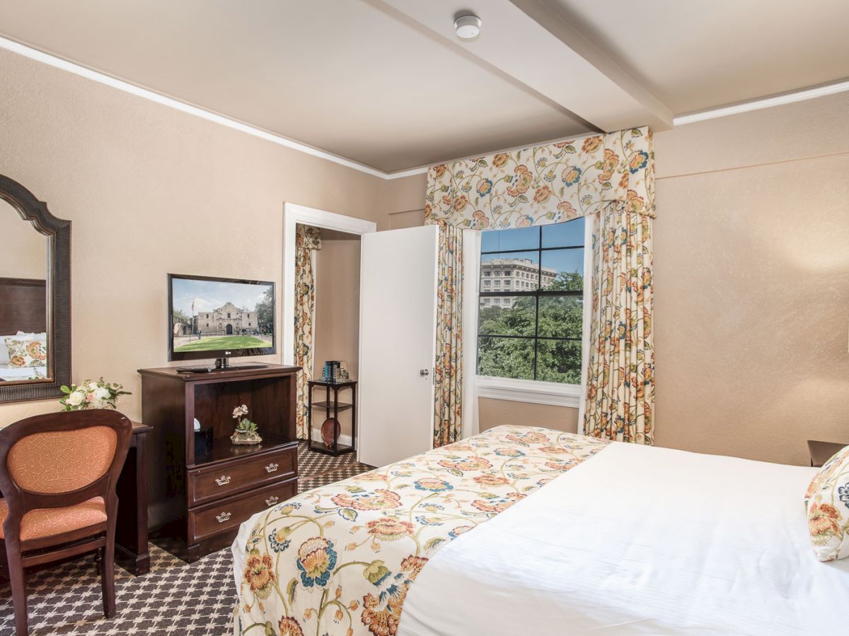 A cozy hotel room with a floral bedspread, dresser, desk with mirror, and a window with floral curtains, inviting workspace and warm lighting.