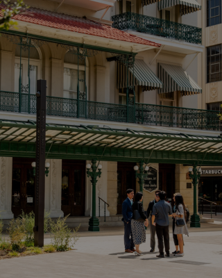 A group of people stands and chats outside a stylish building with striped awnings and a Starbucks, in a plaza setting.
