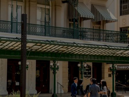 A sunny plaza in front of a stylish building with green balconies and striped awnings; a small group chats near a Starbucks.