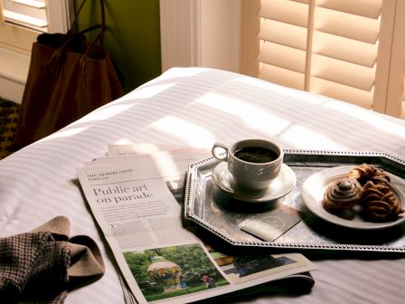 Breakfast scene: newspaper and pastries on a tray beside a cup of coffee, with sun through window blinds.