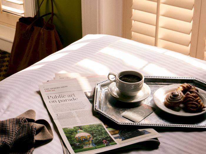 Breakfast scene: newspaper and pastries on a tray beside a cup of coffee, with sun through window blinds.
