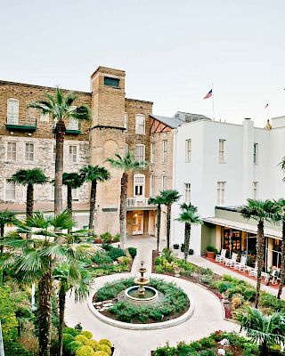 A charming courtyard with a round fountain, palm trees, and surrounding buildings; a sunny, upscale plaza vibe. End.
