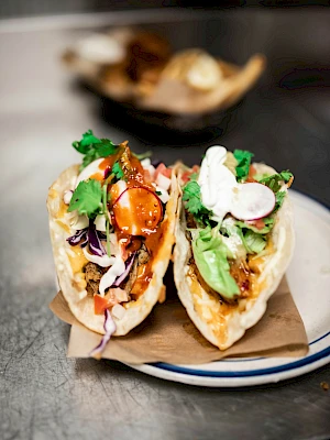 Two crispy taco boats filled with colorful veggies, herbs, dollops of sauce, and a drizzle of chili glaze on top, served on a plate.