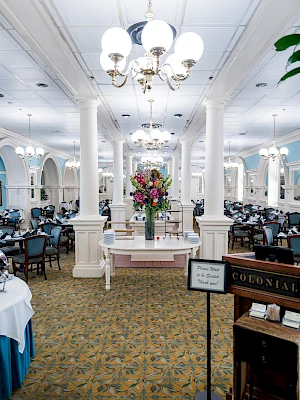 A grand, bright dining hall with ornate white columns, chandeliers, floral centerpiece, and elegantly set tables on a patterned carpet.
