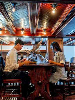 A man and a woman sit across from each other at a wooden bar, sharing a meal in a cozy, warmly lit restaurant with a mounted deer centerpiece.