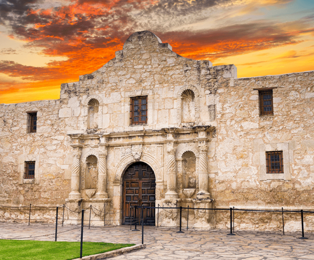Historic stone mission facade with arched doorway, two side windows, a central pediment, and a vibrant sunset sky behind it.