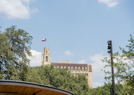 A tall, historic building rises behind trees, with a small flag fluttering atop it, a traffic light on the right, and a curved roof in the foreground.
