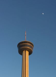 A tall observation tower with a circular platform, clean blue sky, and a small moon visible above.