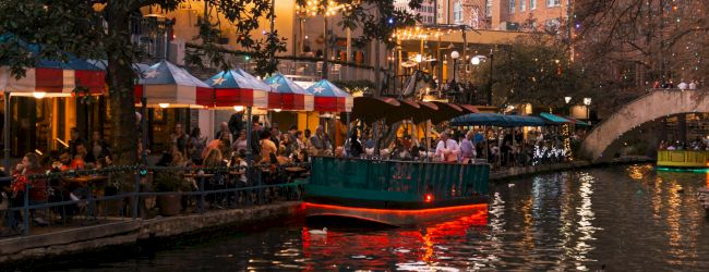 People dine along a canal at sunset, a boat glides by, colorful tents line the shore, warm lights reflect on gently rippling water.