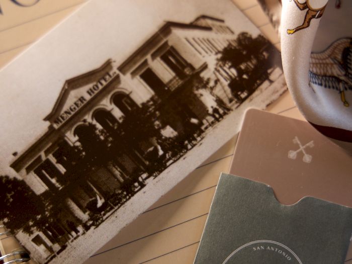 A Menger Hotel package sits beside a notebook with a vintage &ldquo;Menger Hotel&rdquo; photo postcard and a decorated bookmark.