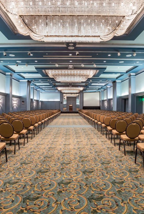 A large conference hall set up with rows of chairs facing a stage, chandeliers overhead, and patterned carpet, ready for a ceremony or presentation.