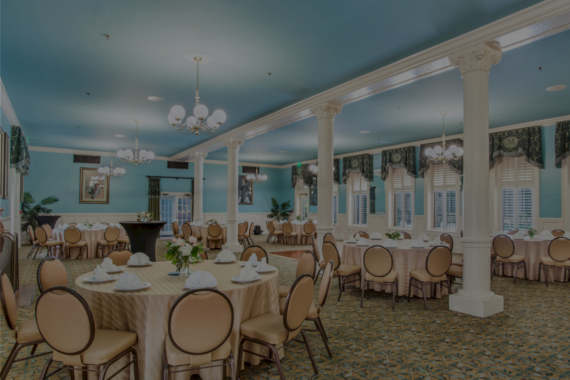 A ballroom set for a banquet with round tables, cream tablecloths, beige chairs, chandeliers, columns, and blue ceiling in a bright, elegant dining hall.