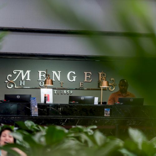 A small outdoor cafe or storefront with a sign, greenery in front, and a person seated at a table; text is partially legible.