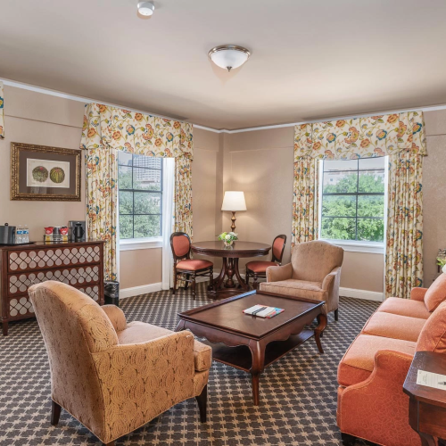 A cozy living room with floral curtains, beige walls, and a mix of upholstered chairs and a sofa around a wooden coffee table, sunny windows, and lamps.