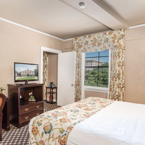 A cozy hotel room with a floral bedspread, vanity mirror, dresser, desk, chair, patterned curtains, and a window view of greenery, warmly lit.